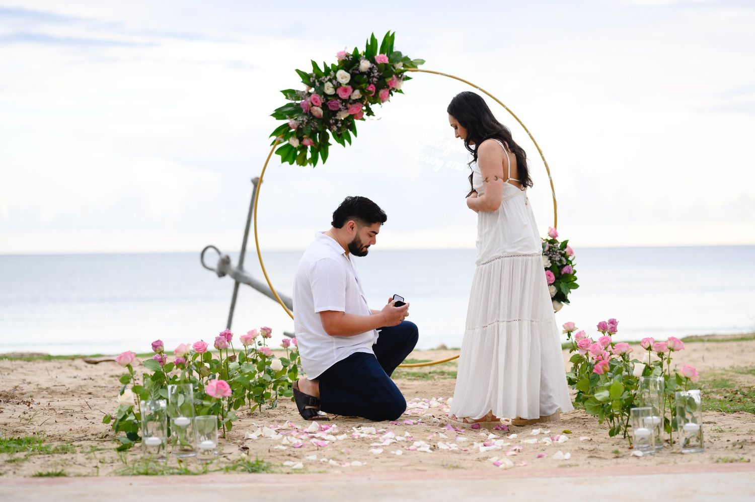 Man proposing to woman on beach under floral arch with roses, romantic ocean backdrop.