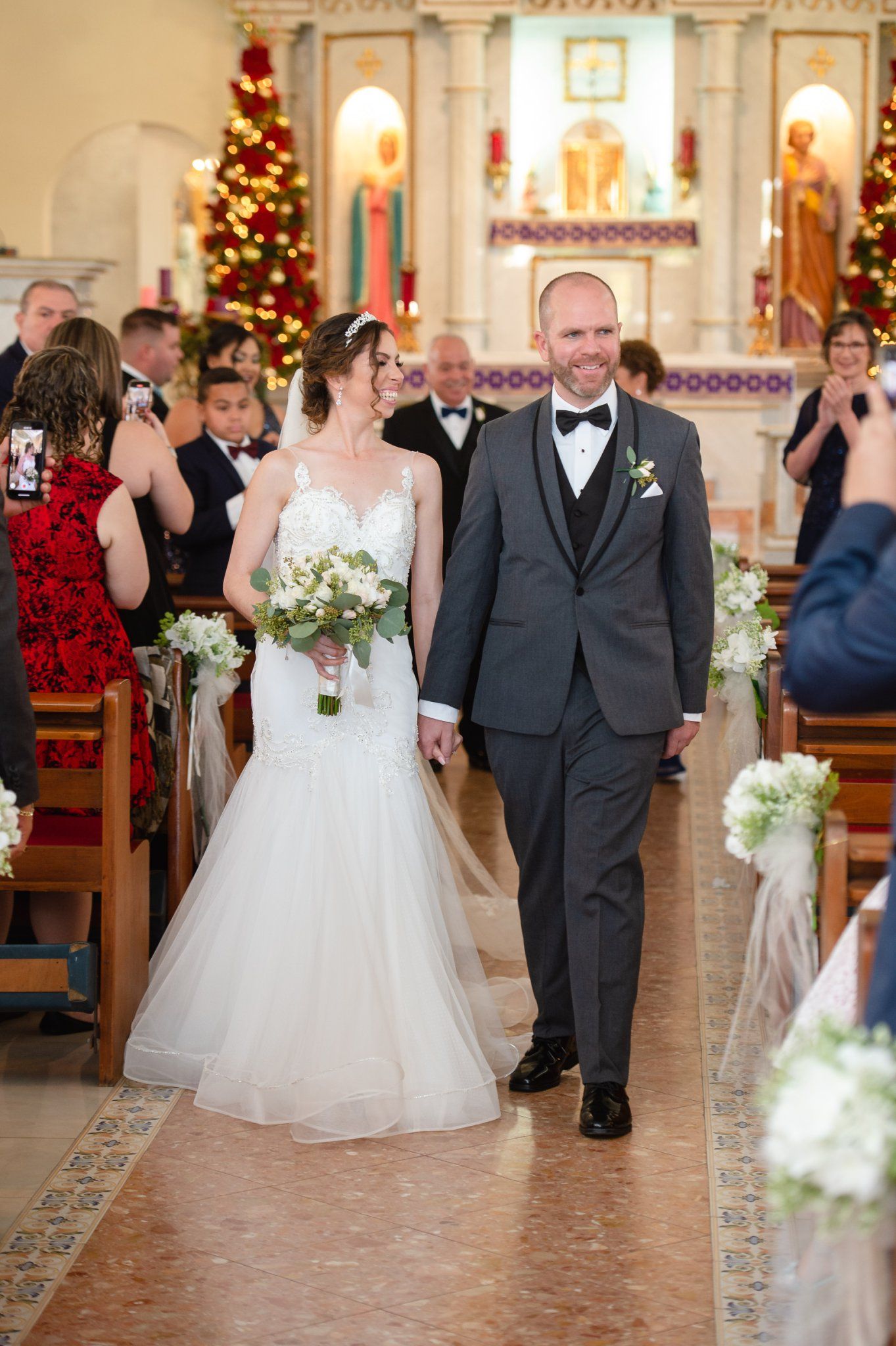 Bride and groom walk down church aisle after wedding ceremony, decorated with white flowers and Christmas trees.