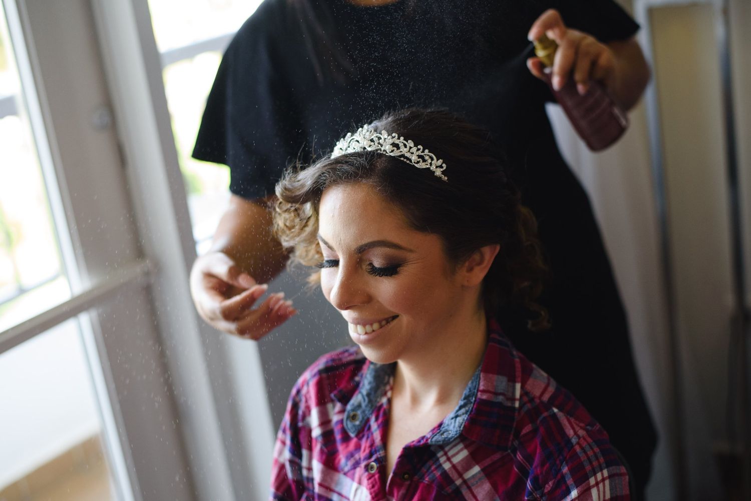 Smiling bride wearing tiara gets hair styled and sprayed before wedding ceremony.
