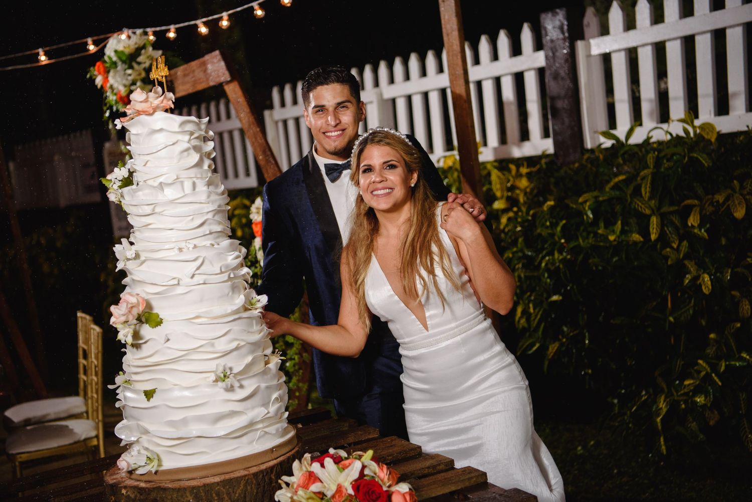 Bride and groom smiling beside a tall white tiered wedding cake decorated with flowers at an outdoor night reception.