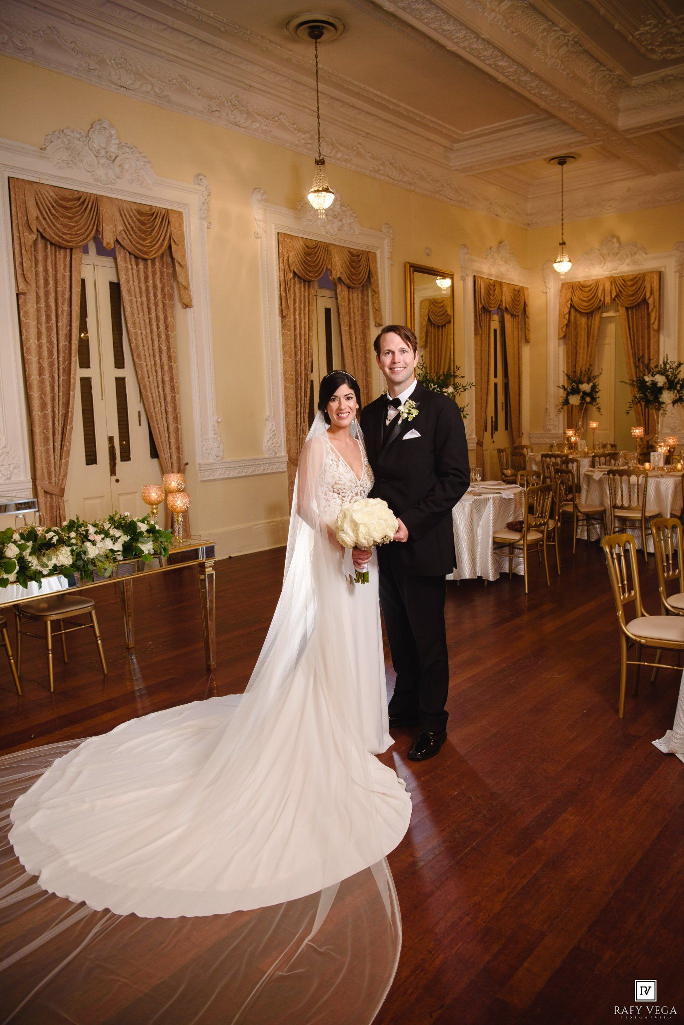 Bride in white gown with long veil and groom in black tuxedo pose together in elegant ballroom wedding venue.