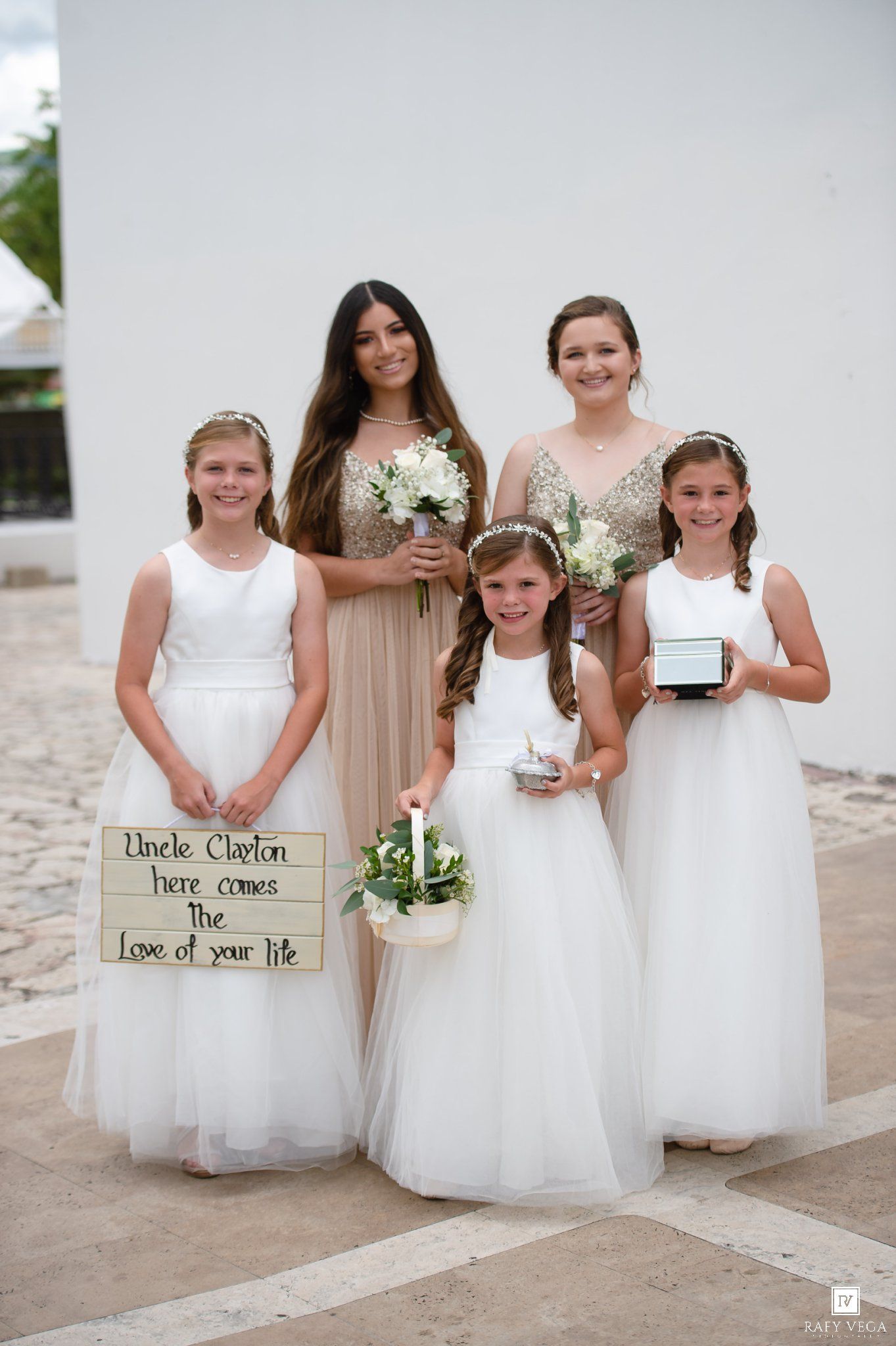 Flower girls and bridesmaid in white dresses pose with sign reading Uncle Clayton here comes the love of your life.