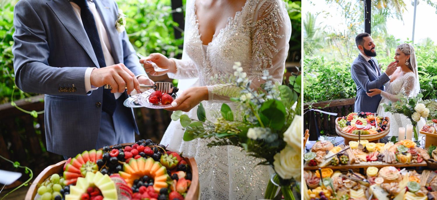 Bride and groom cutting wedding cake beside elegant fruit and charcuterie grazing tables at outdoor reception.