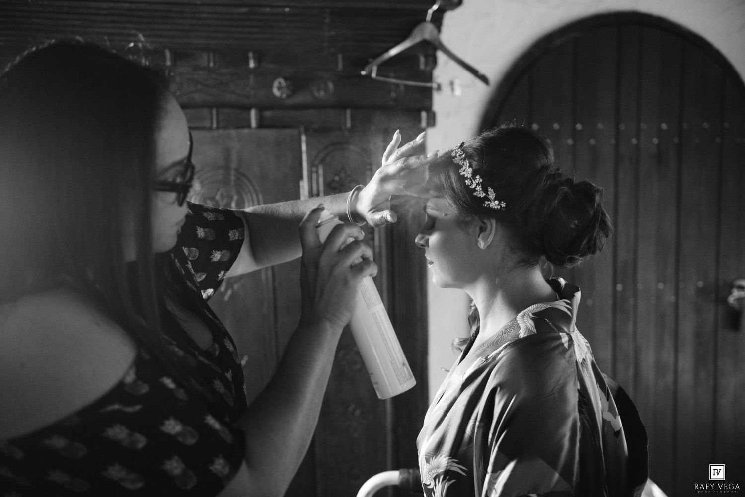 Black and white photo of a bride getting her hair styled by a makeup artist wearing a face mask before her wedding.