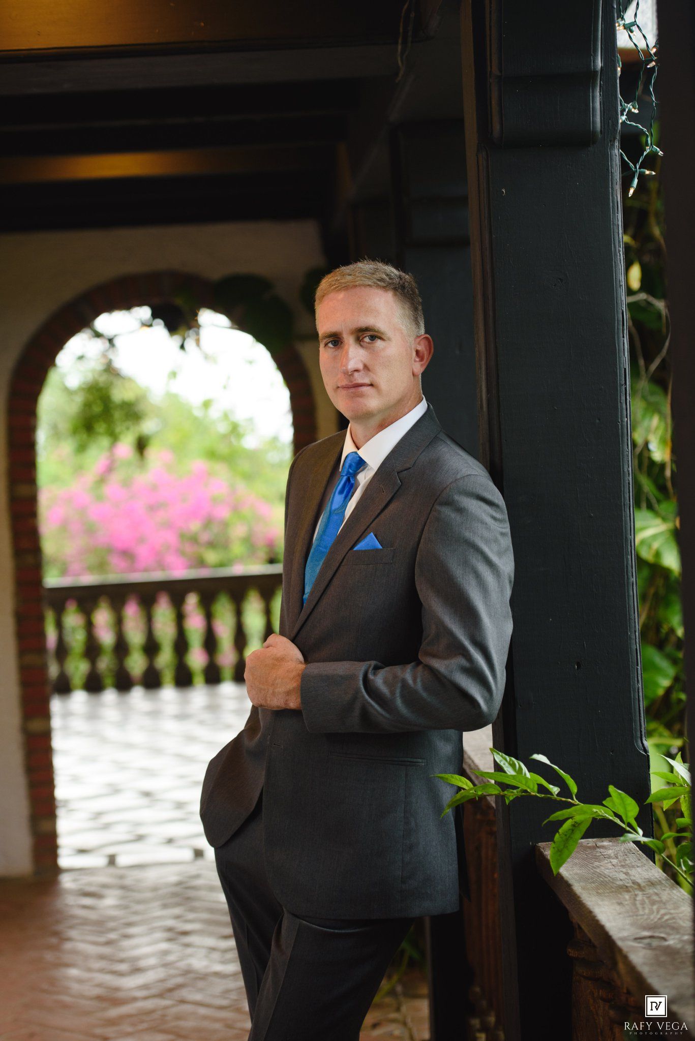 Man in gray suit with blue tie and pocket square poses elegantly on a colonial-style terrace with lush greenery.