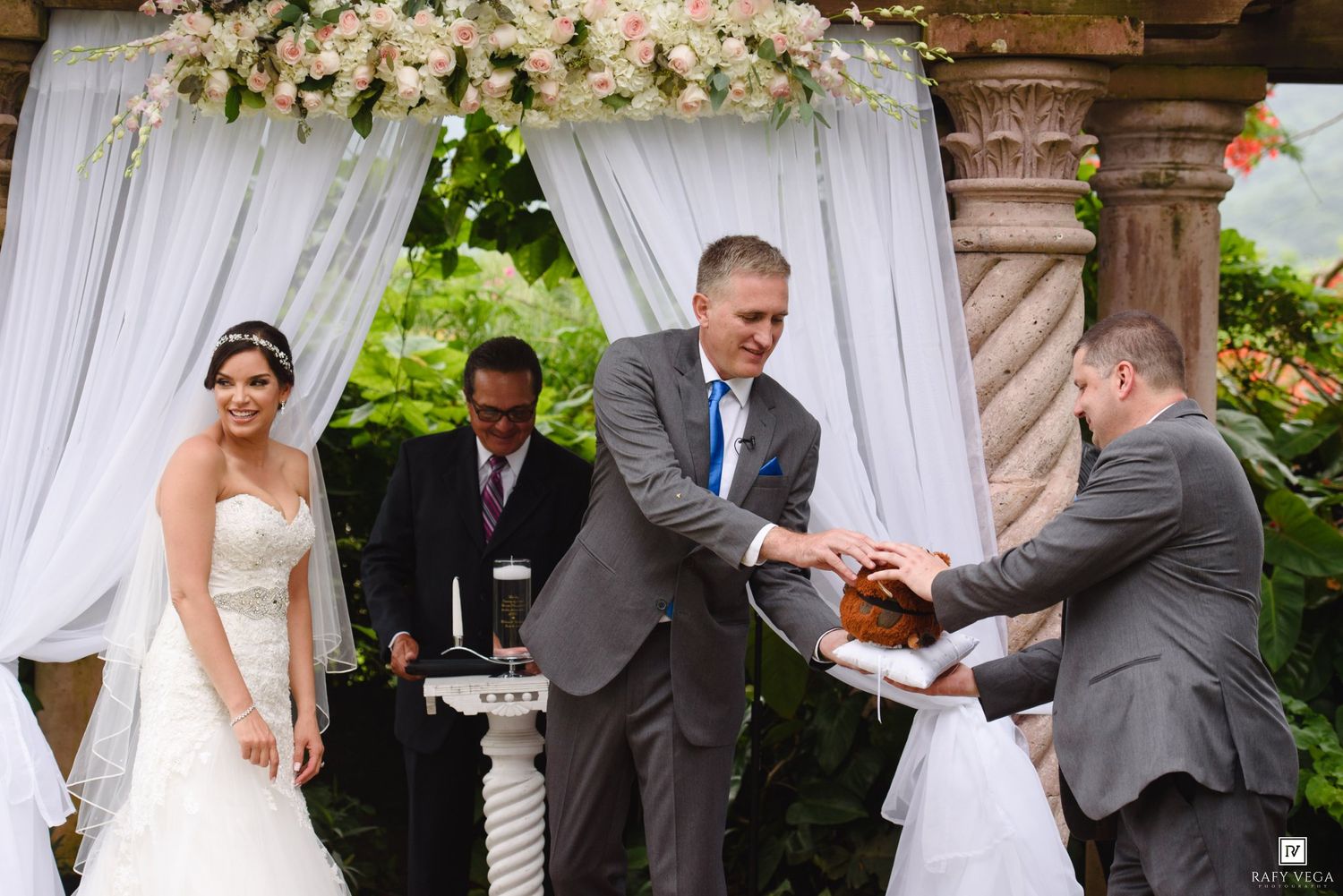 Bride and groom exchanging rings at an elegant outdoor wedding ceremony under a floral arch with draped white fabric.