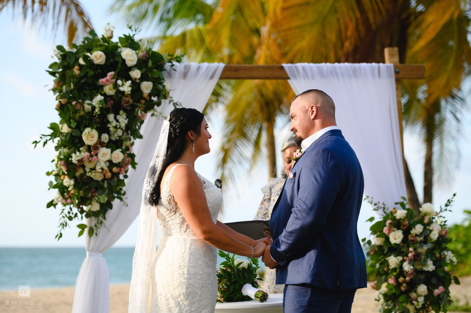Bride and groom hold hands at beachside altar adorned with lush floral arch during outdoor wedding ceremony.