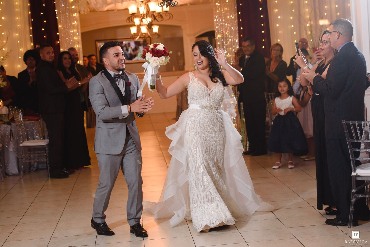 Bride in white lace gown and groom in gray suit share first dance at elegant wedding reception with fairy lights.
