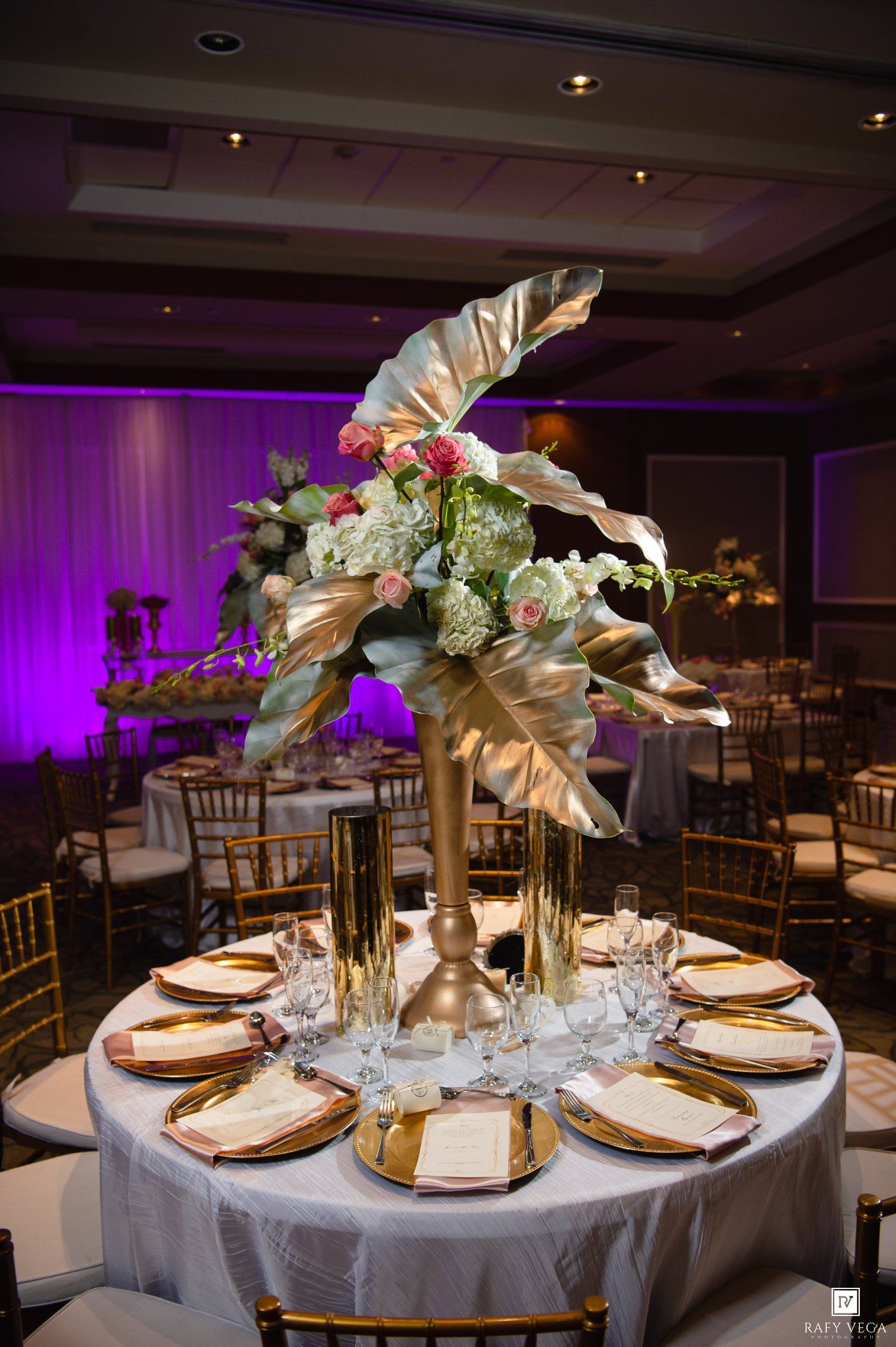 Elegant wedding reception table with gold floral centerpiece, white linens, and gold charger plates in a ballroom with purple uplighting.