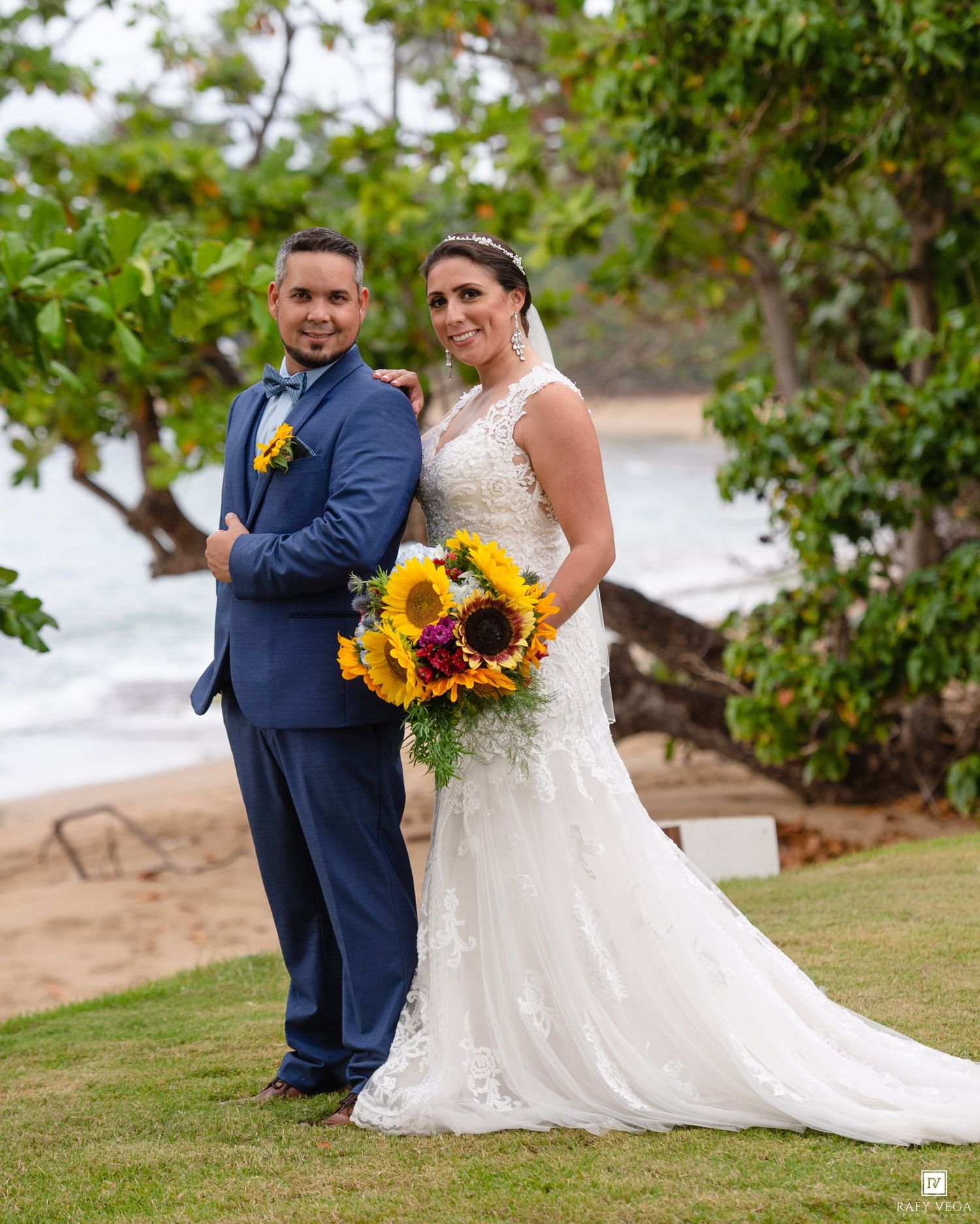 Bride and groom pose on beach lawn, she holds sunflower bouquet in white lace gown, he wears navy suit.
