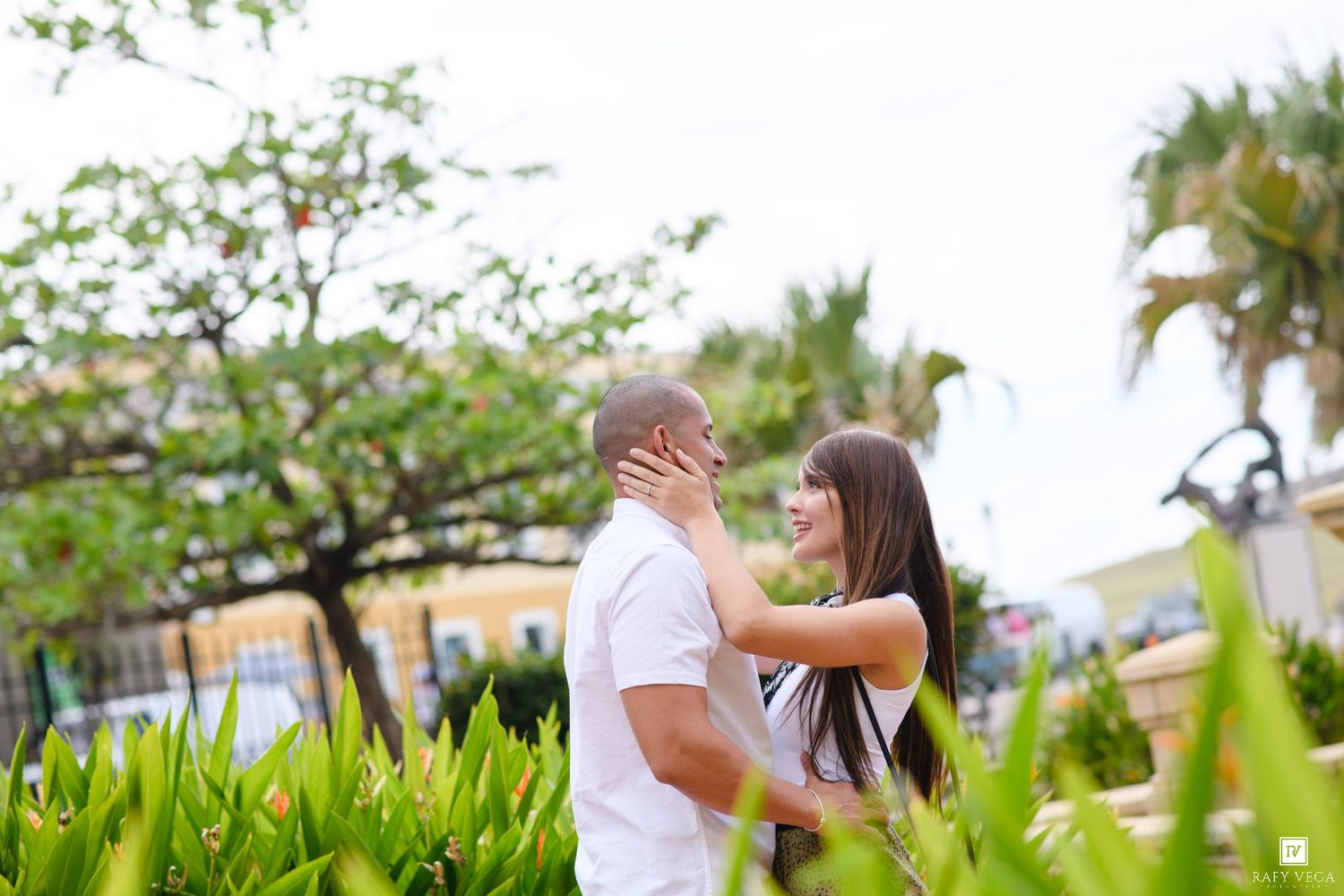 Romantic couple embracing outdoors among tropical greenery, smiling at each other in a sunny garden setting.