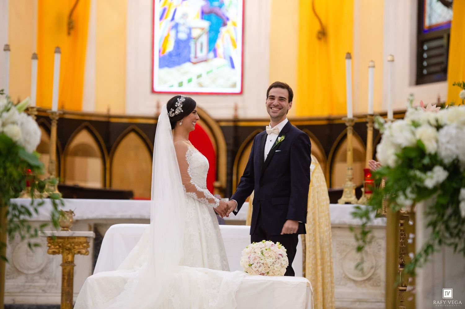 Bride and groom holding hands at church altar during wedding ceremony with floral arrangements.