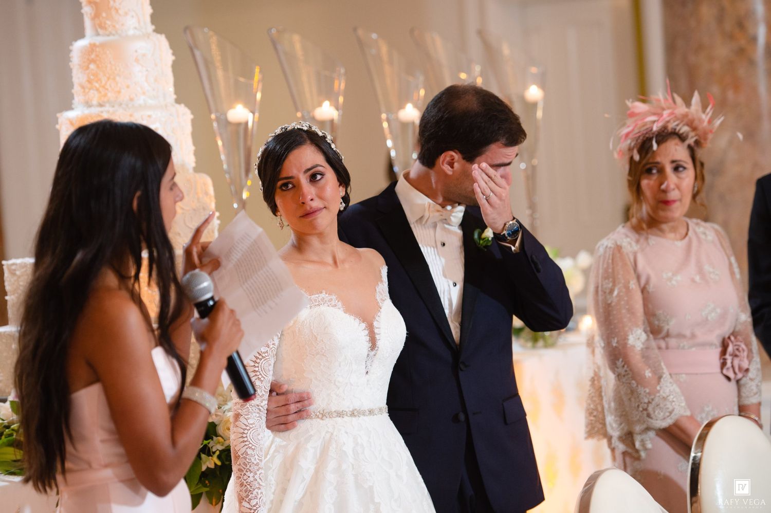 Emotional groom covers mouth as bride listens to speech near wedding cake at reception.