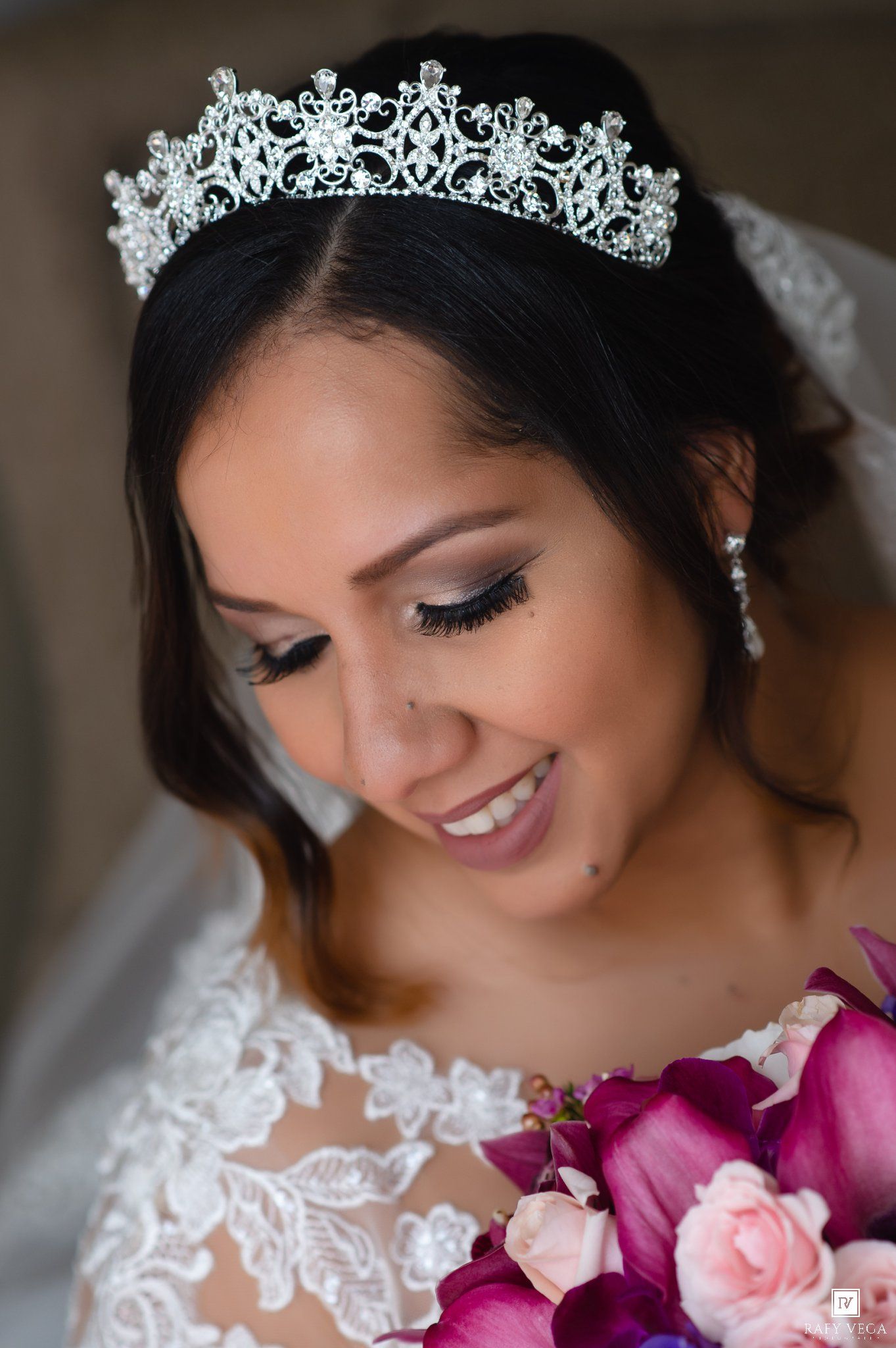 Smiling bride wearing a sparkling tiara and lace gown holds a bouquet of pink and purple flowers on her wedding day.