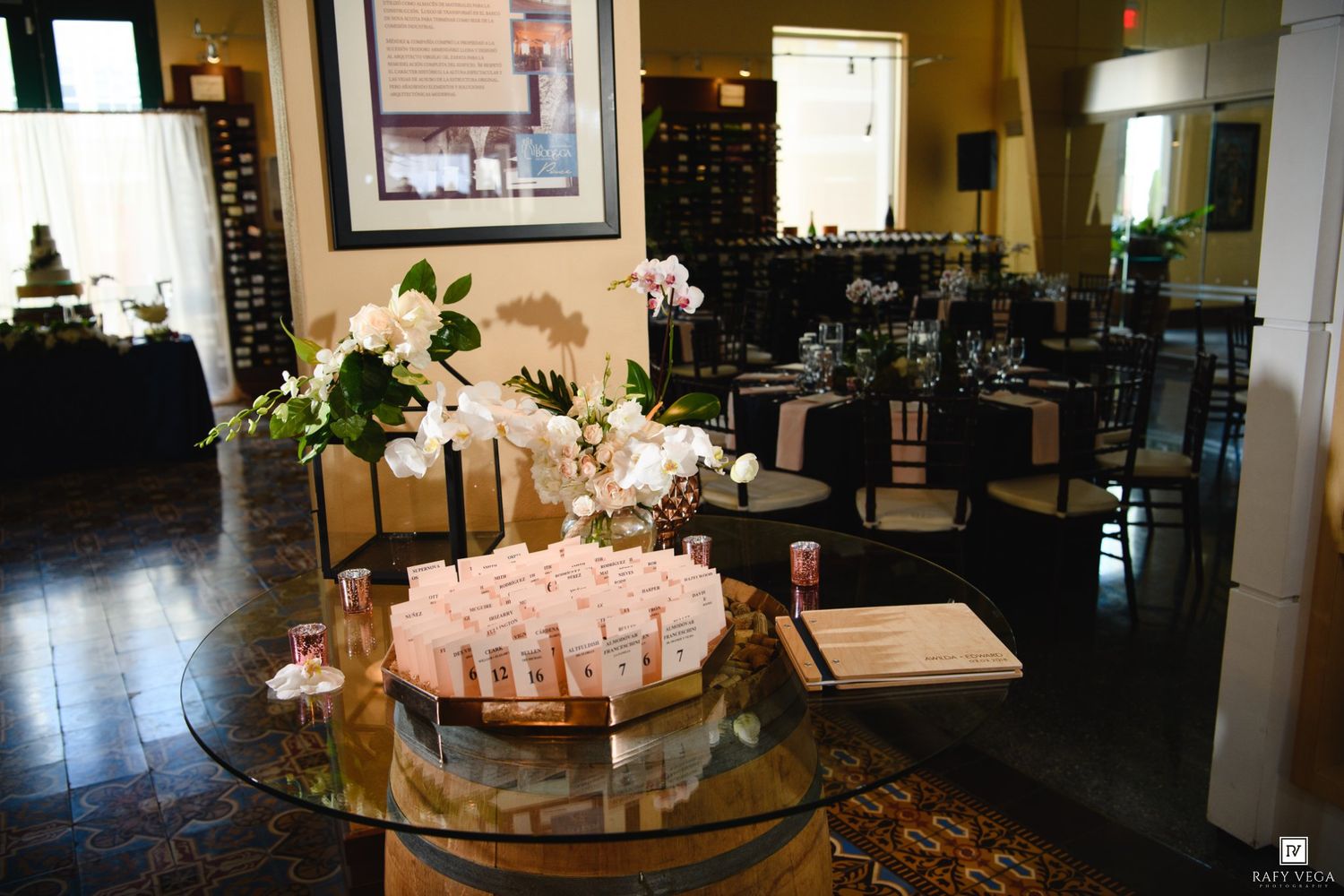 Elegant event venue with glass table displaying floral arrangements and place cards, dark wood floors, and dining tables in background.