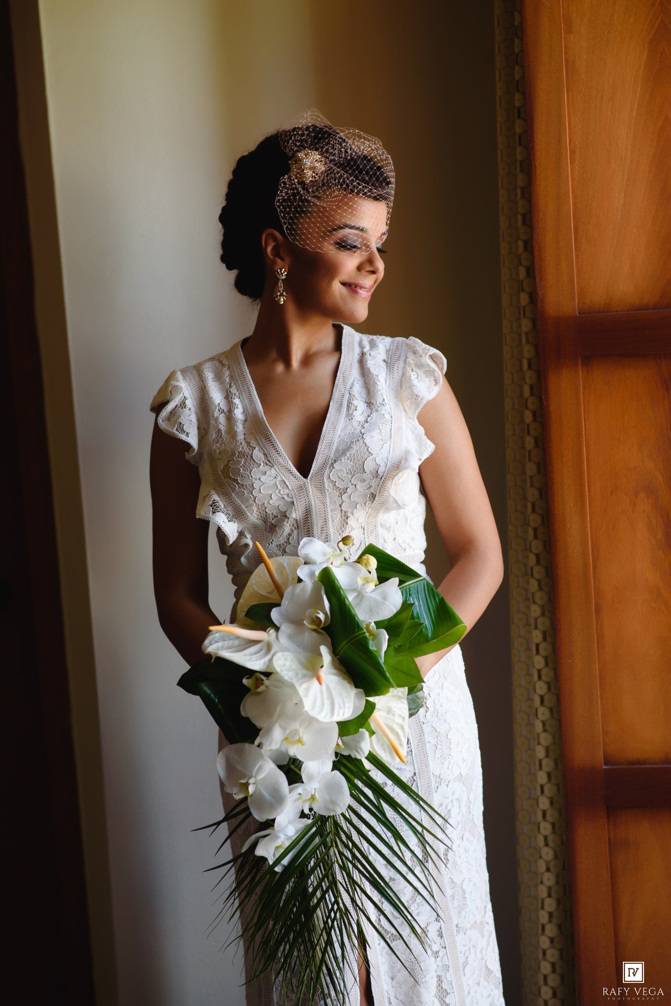 Elegant bride in white lace dress holding tropical bouquet with orchids and palm leaves, standing by a window.