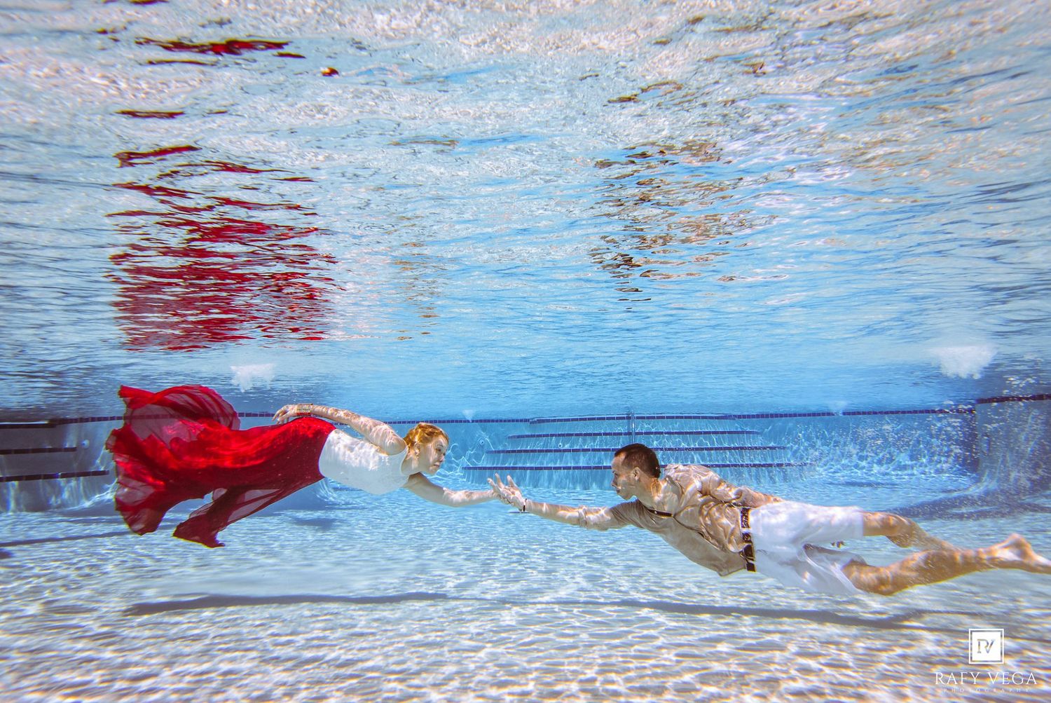 Couple posing underwater in a pool, woman in flowing red dress and man in casual attire, holding hands gracefully.