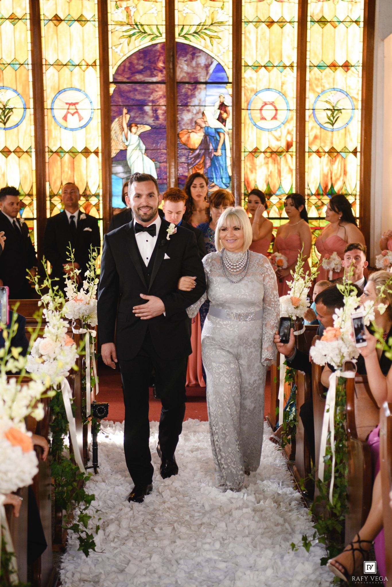 Groom walks down church aisle with mother and child before stained glass window at elegant wedding ceremony.