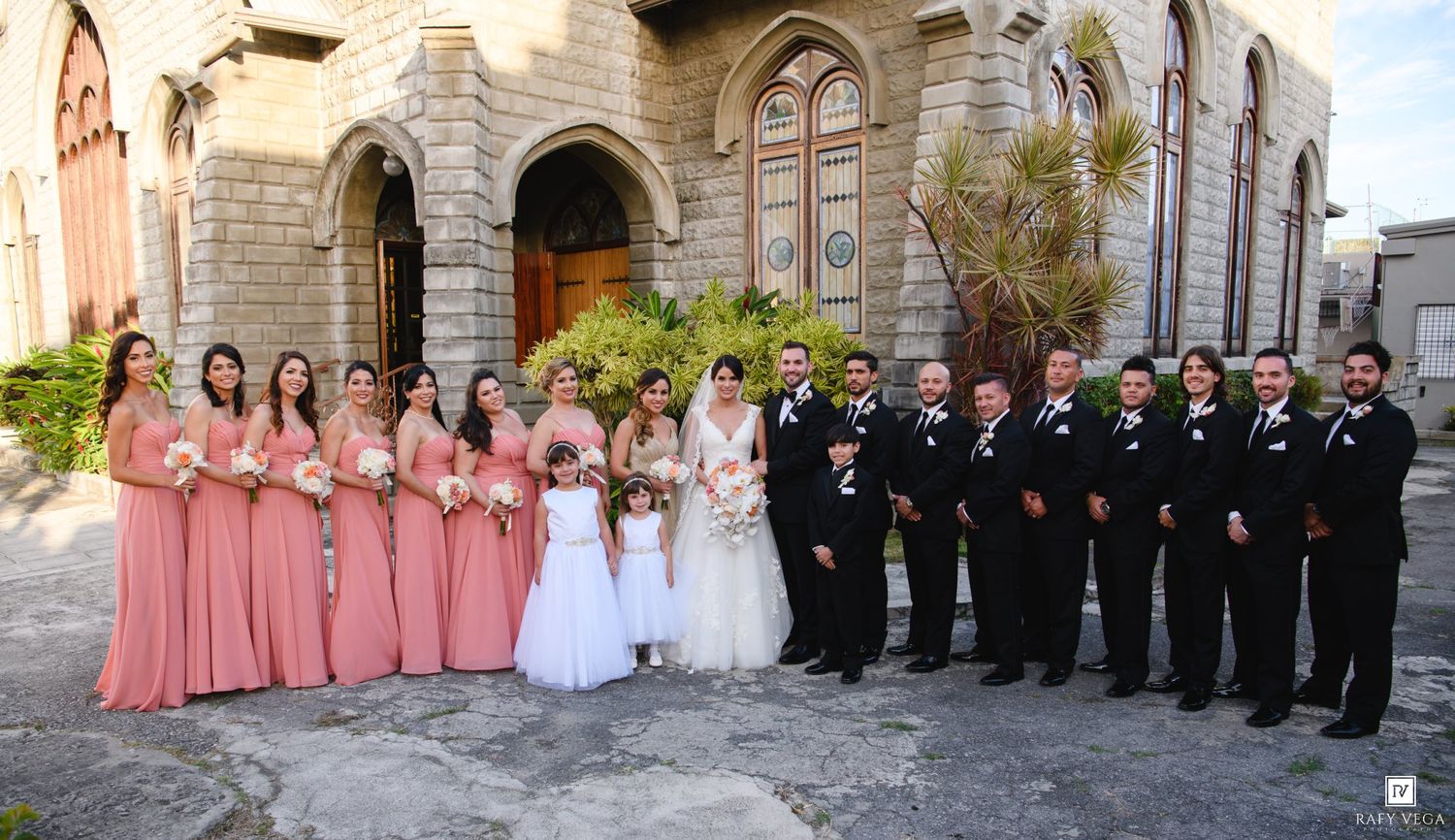 Wedding party posing outside stone church, bridesmaids in coral gowns and groomsmen in black suits flanking bride and groom.