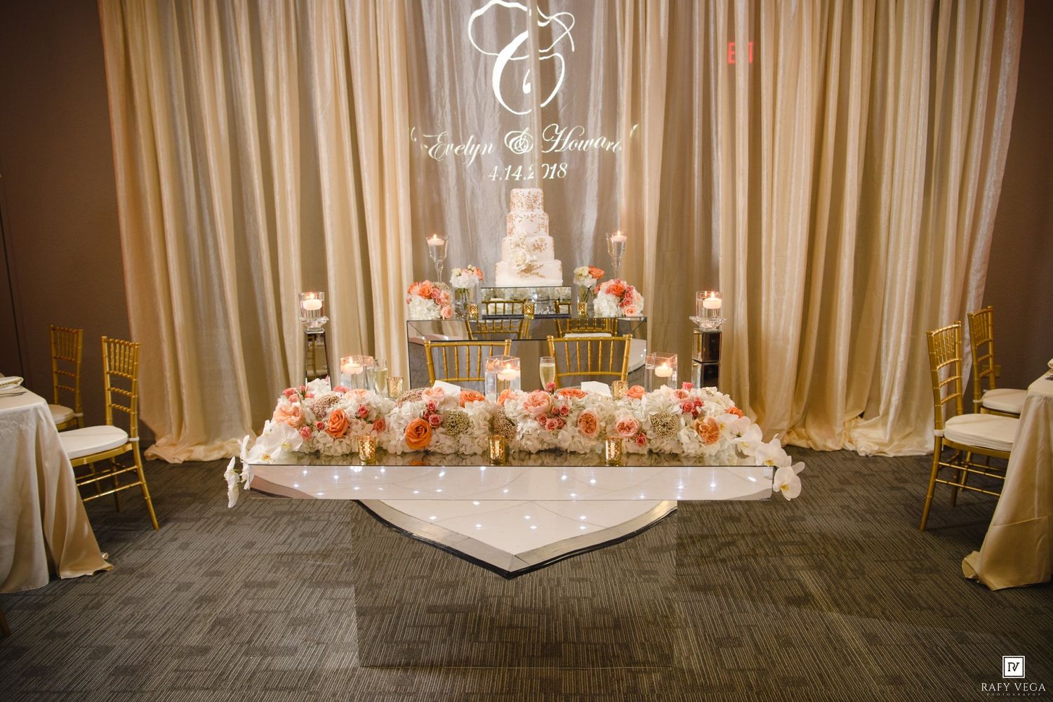 Elegant wedding reception sweetheart table with peach floral arrangements, gold chairs, candles, and monogram backdrop.