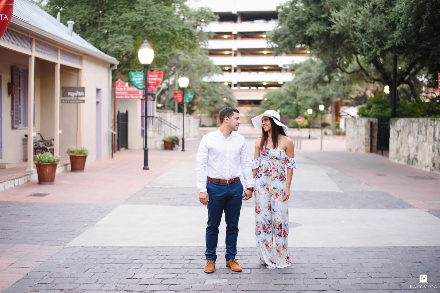 Couple holding hands on a brick walkway in a charming outdoor shopping district, woman in floral maxi dress and sun hat.