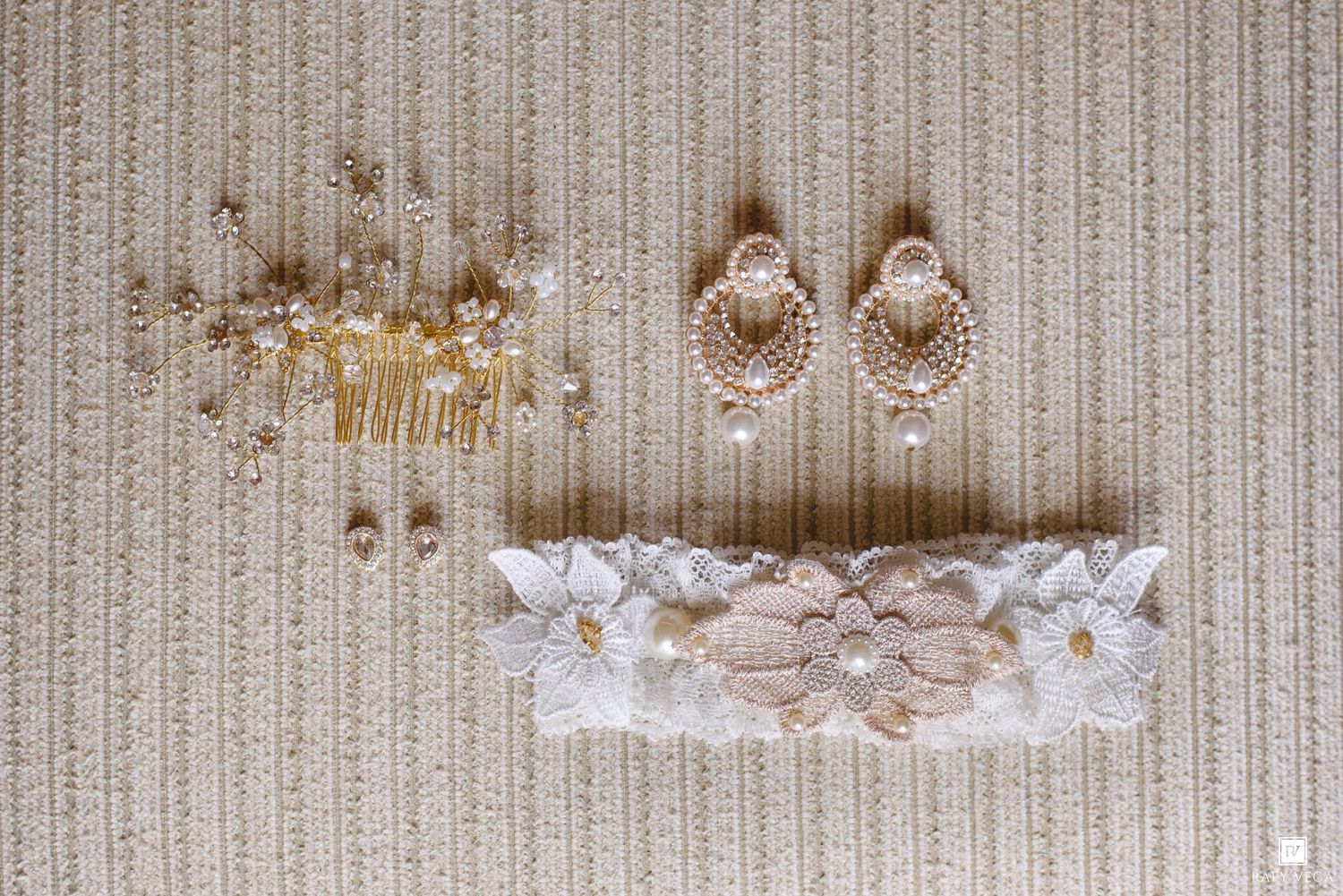 Bridal accessories flat lay featuring gold hair comb, pearl drop earrings, and white lace garter on beige fabric.