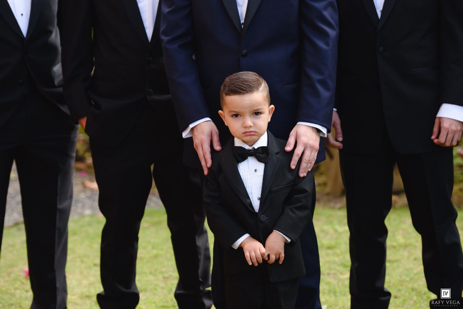 Young boy in tuxedo stands proudly with groomsmen in suits at a wedding ceremony outdoors.