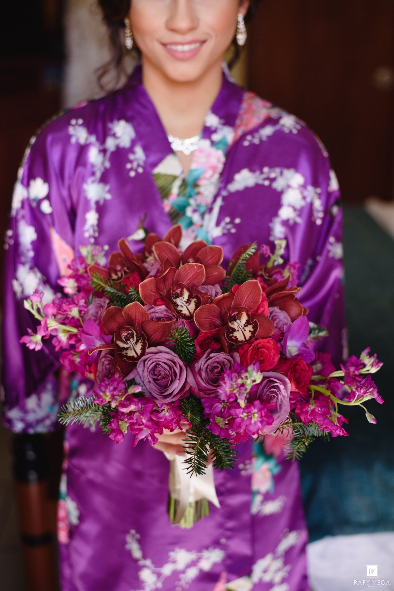 Bride in purple floral satin robe holding a lush bouquet of red orchids, magenta roses, and deep purple blooms.