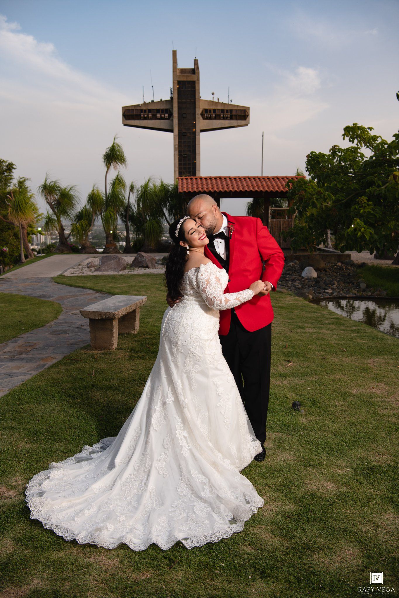 Bride in white lace gown and groom in red tuxedo embrace near a large cross monument on lush green grounds.