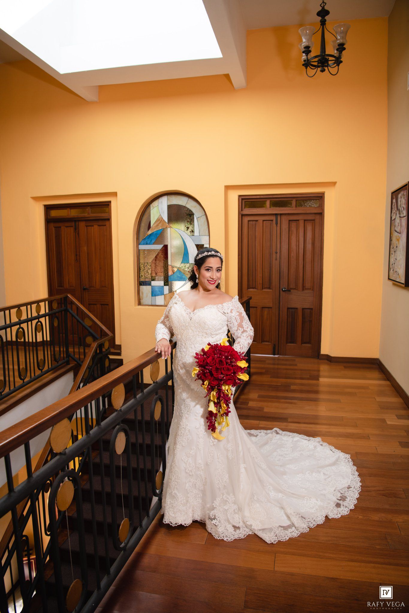 Elegant bride in long-sleeve lace mermaid gown holds red bouquet on wooden staircase landing in yellow-walled venue.