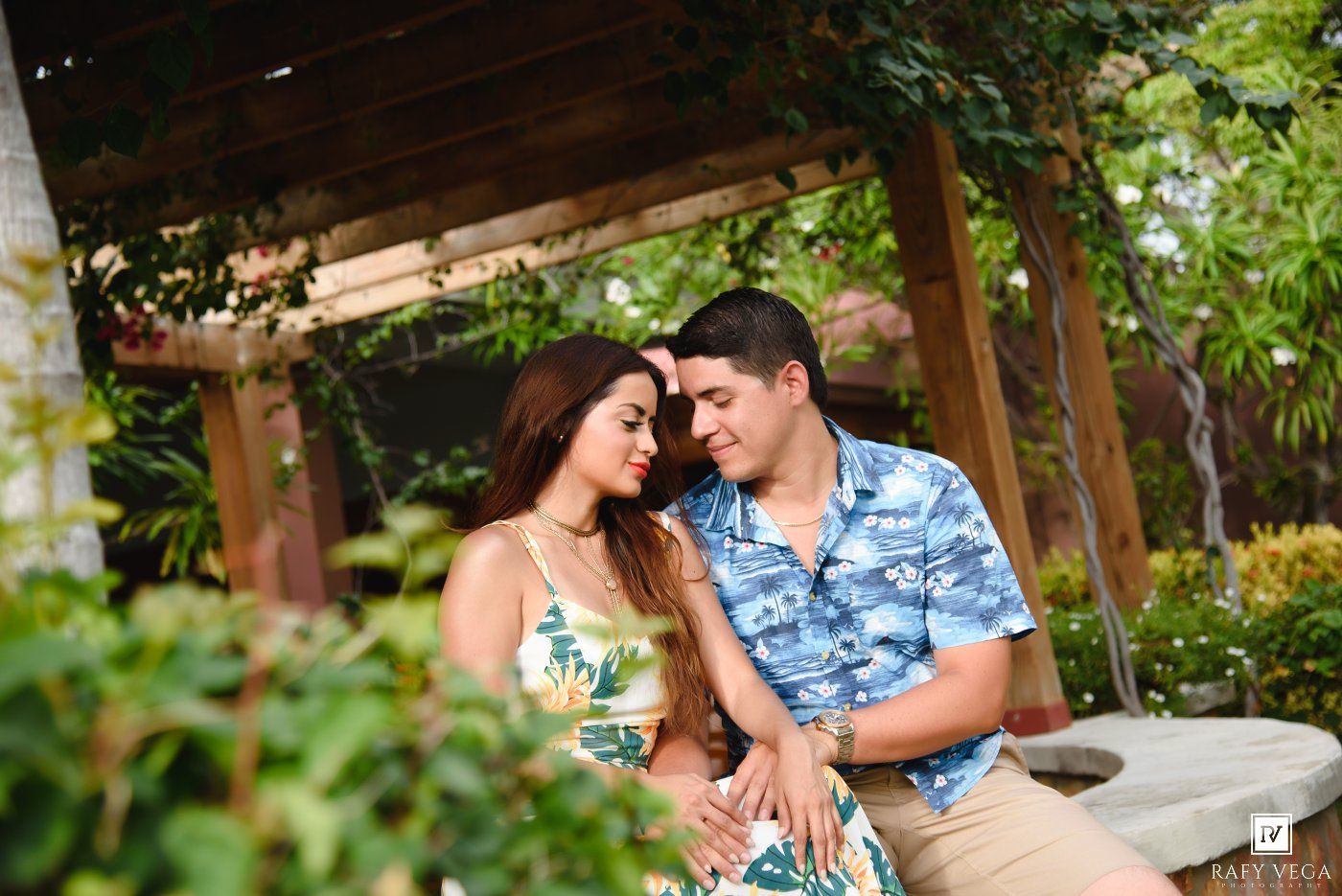 A couple shares a romantic moment sitting together in a lush garden gazebo, wearing tropical attire.