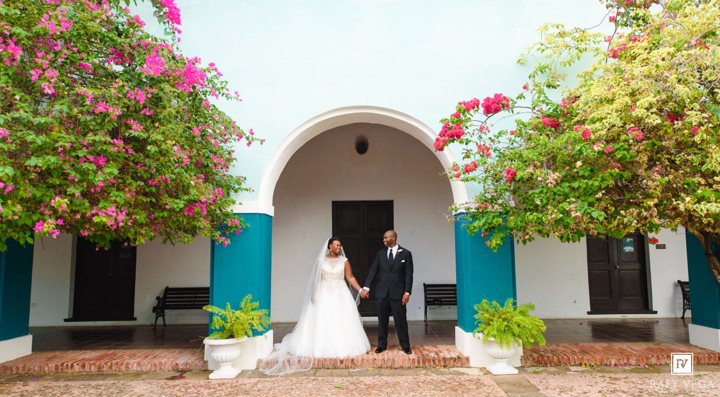 Bride and groom holding hands under a white archway surrounded by vibrant bougainvillea at an outdoor wedding venue.