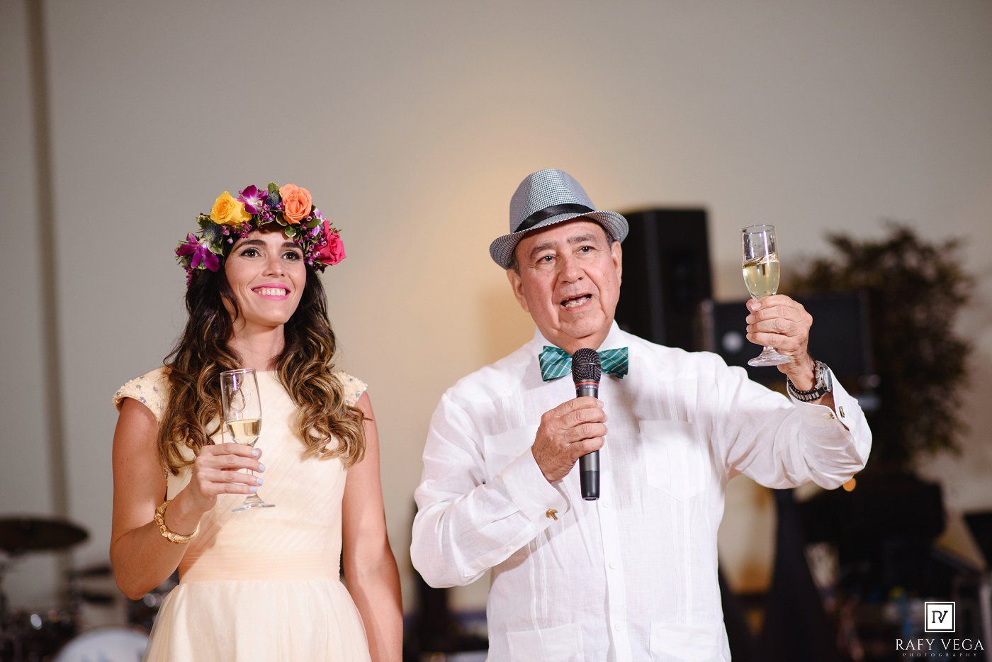 A man in a gray hat and bow tie holds a microphone and champagne glass next to a woman wearing a floral crown at a wedding.