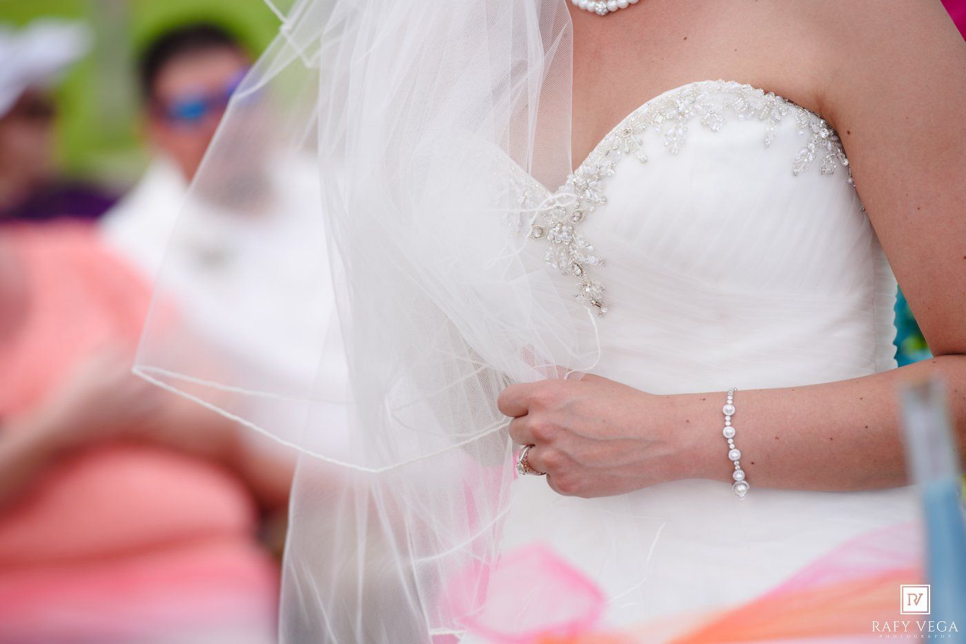 Bride in a strapless white wedding gown with beaded bodice and veil, wearing a delicate bracelet outdoors.