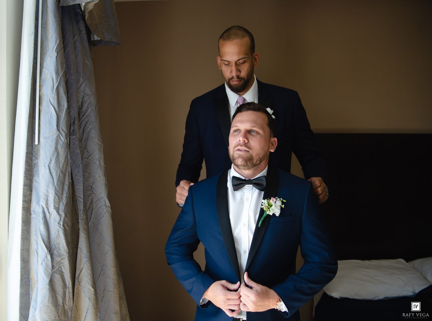 Groom in blue tuxedo gets help adjusting jacket from groomsman before wedding ceremony indoors.