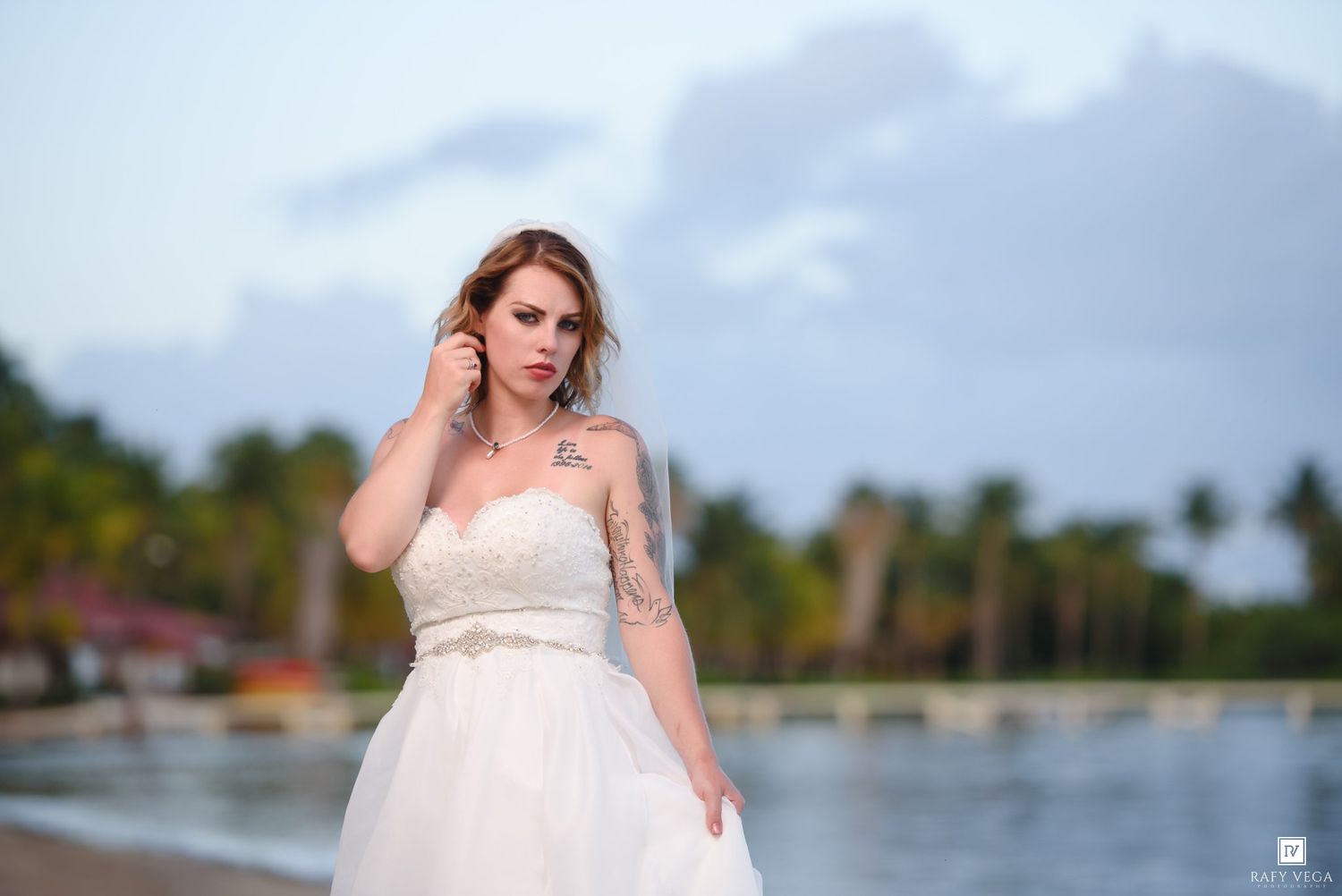 Bride in strapless lace wedding dress posing on tropical beach, touching her ear with palm trees behind her.