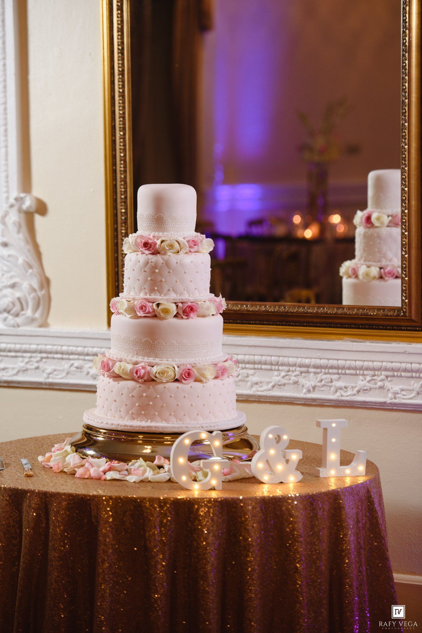 Elegant five-tier white wedding cake adorned with pink roses on a gold stand, displayed in a grand ballroom setting.