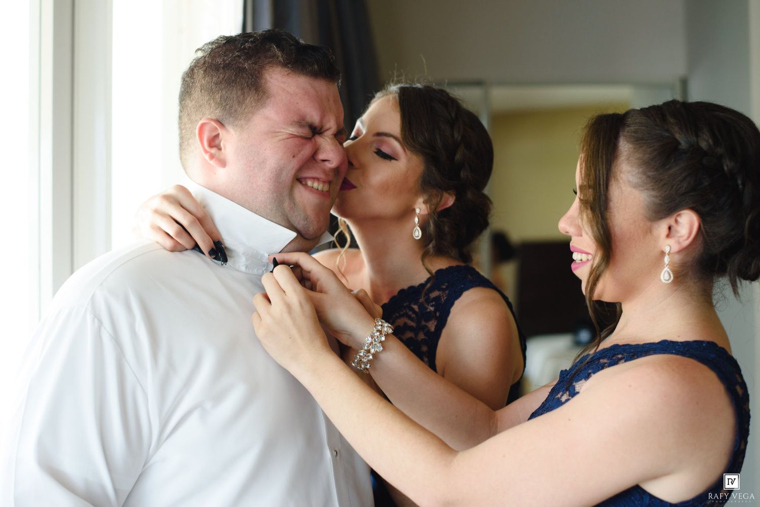 Two women in navy blue dresses help a man adjust his bow tie before a wedding ceremony.