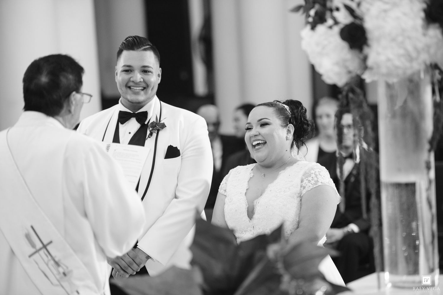Black and white photo of a joyful couple laughing during their wedding ceremony at the altar with an officiant.