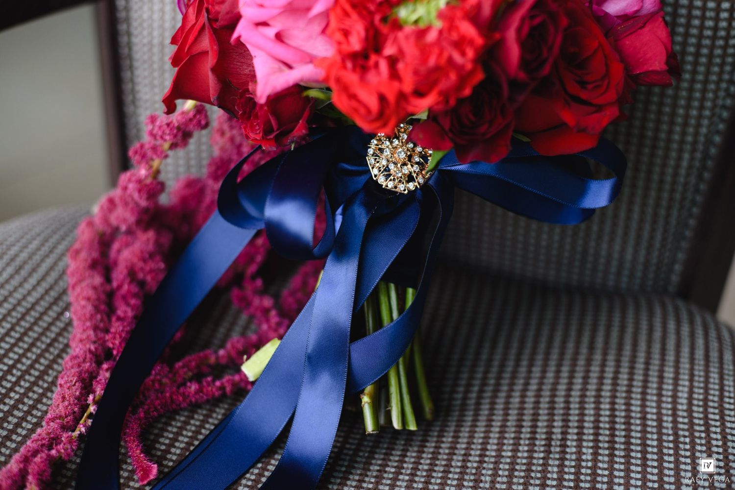 Red and pink bridal bouquet with navy blue satin ribbons and jeweled brooch accent on gray chair.