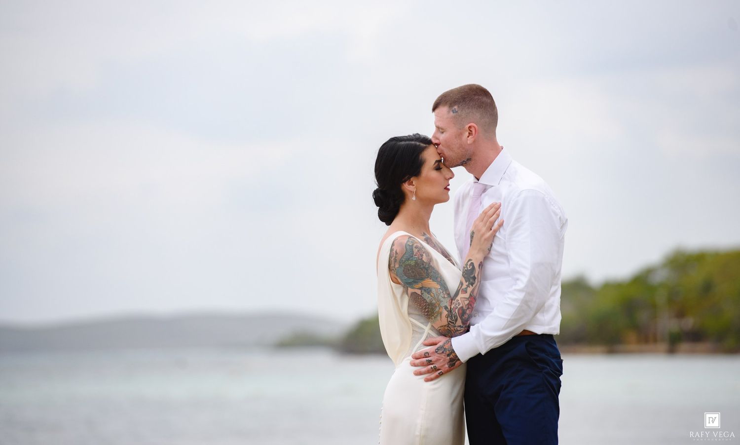 Couple embracing on a beach in wedding attire, groom in white shirt and navy pants, bride with tattoo sleeve in white gown.