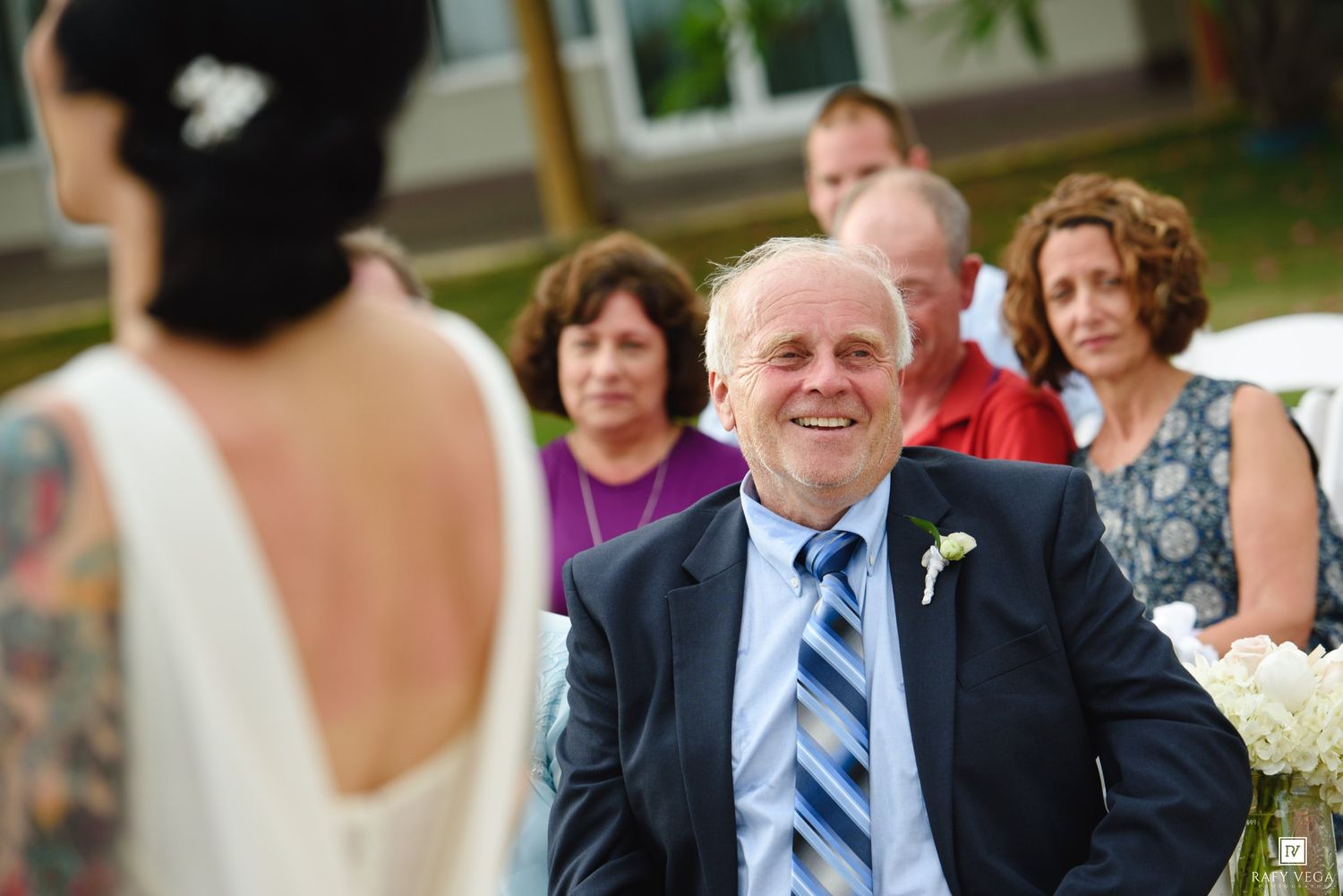 Smiling older man in blue suit with boutonniere watches bride at outdoor wedding ceremony with guests seated behind him.