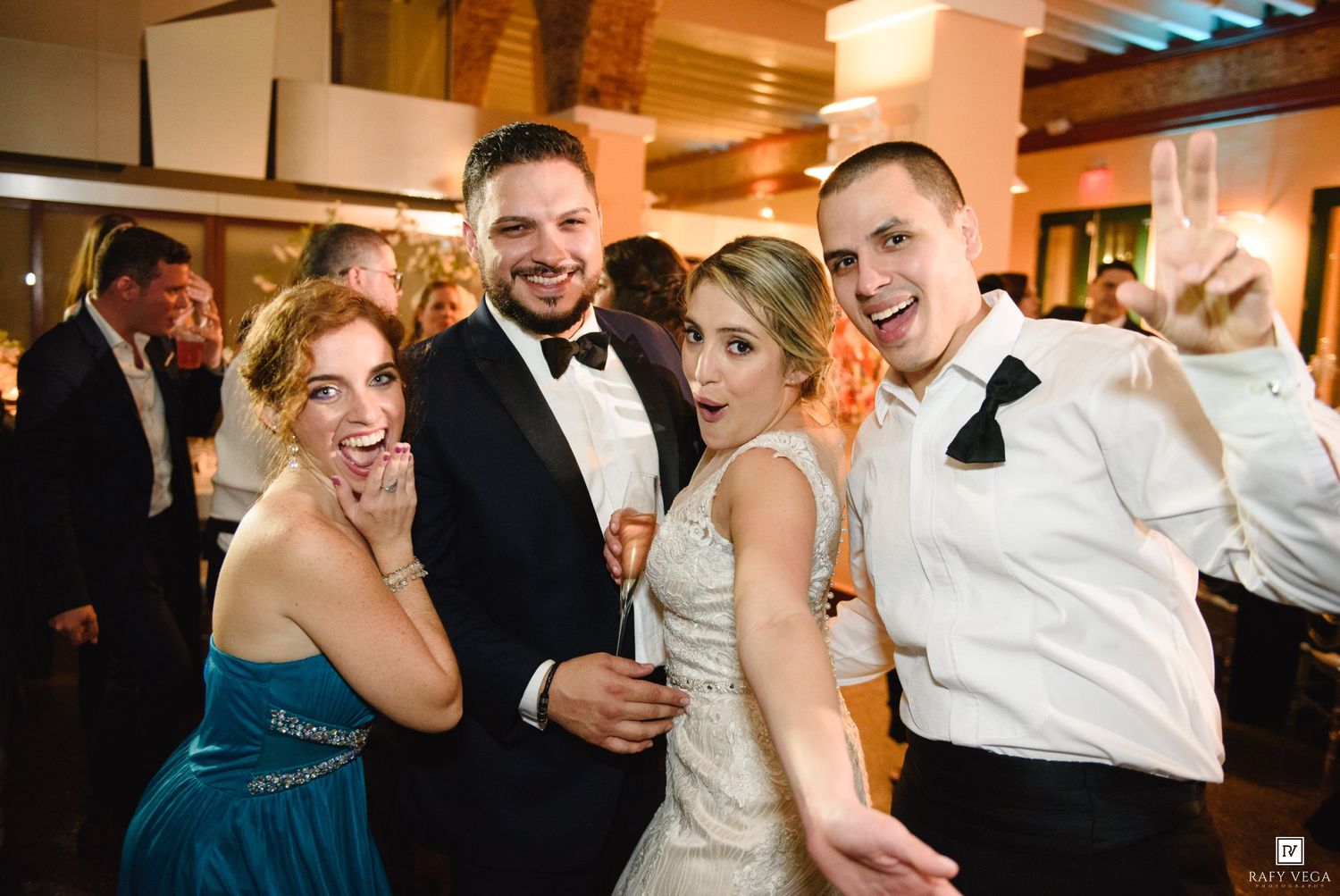 Joyful wedding reception group photo with bride in white gown, groom in tuxedo, and guests celebrating with champagne.