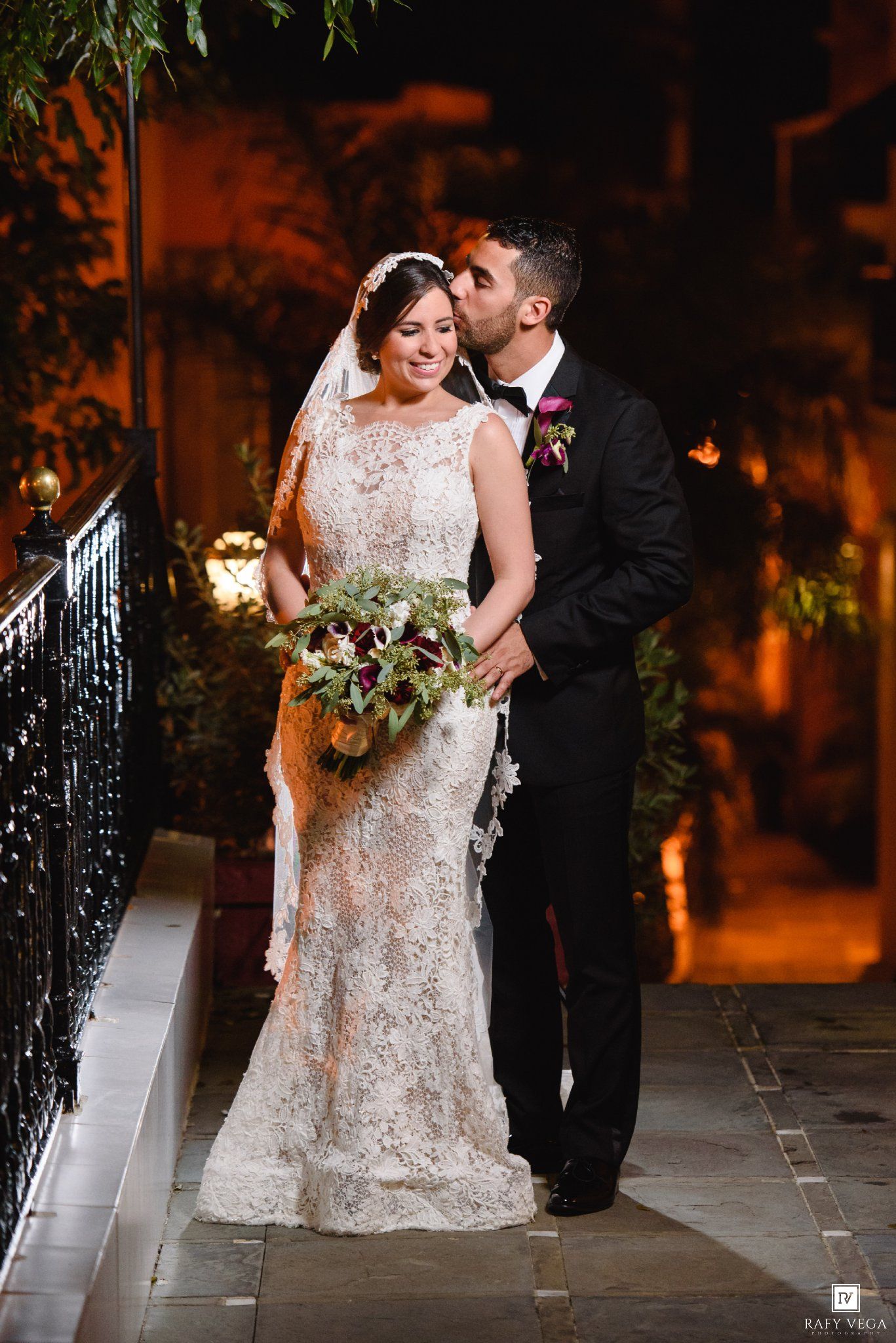 Bride in lace gown holds bouquet as groom in tuxedo kisses her cheek during romantic outdoor evening wedding portrait.