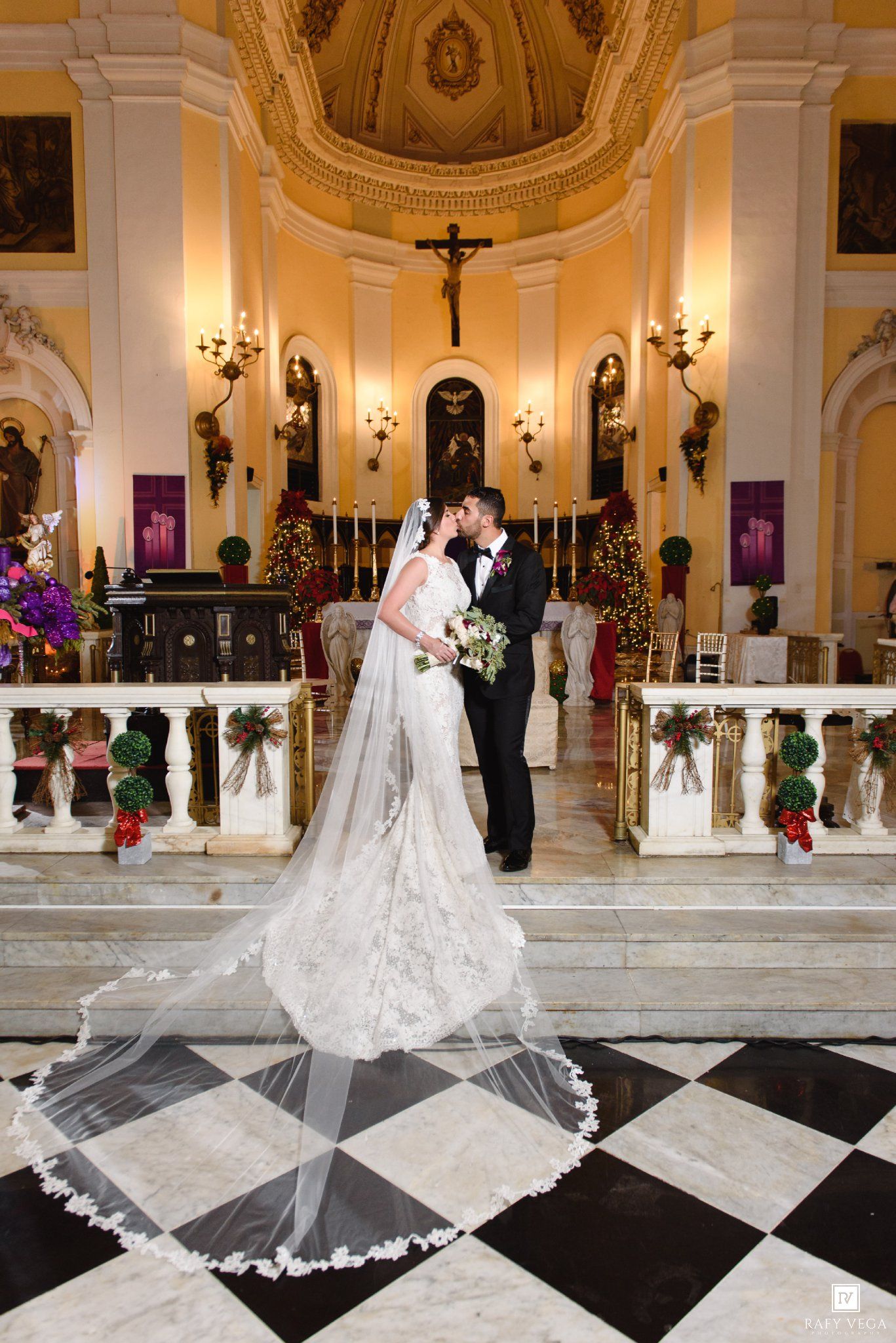 Bride and groom share a kiss at the altar of an ornate church with checkered floors and holiday decorations.