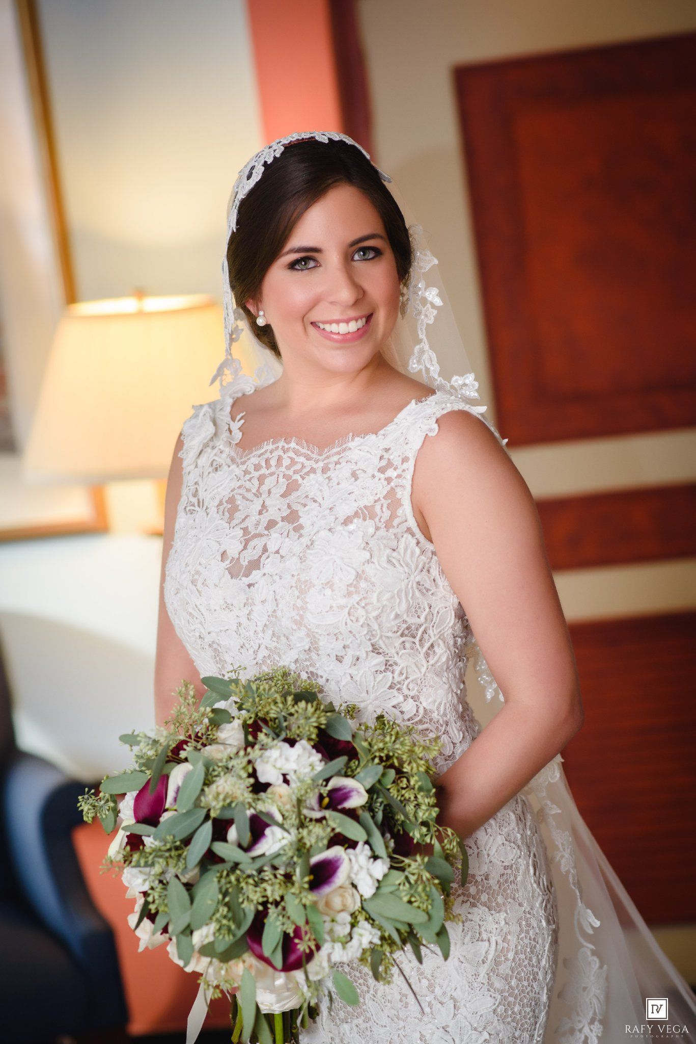 Smiling bride in lace wedding dress holding purple and white bouquet with greenery, wearing veil and tiara.