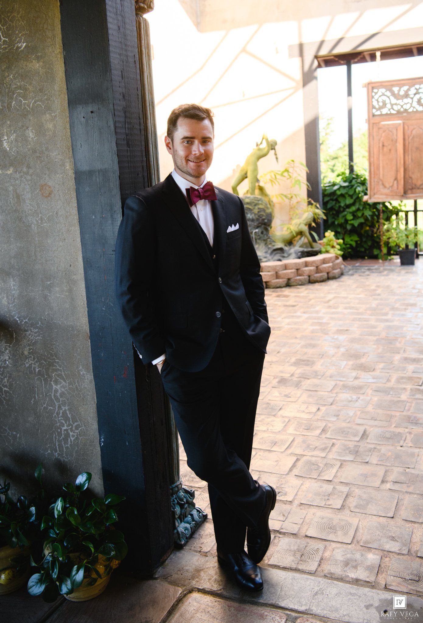 Smiling groom in black tuxedo with burgundy bow tie leaning against a pillar at an outdoor wedding venue.