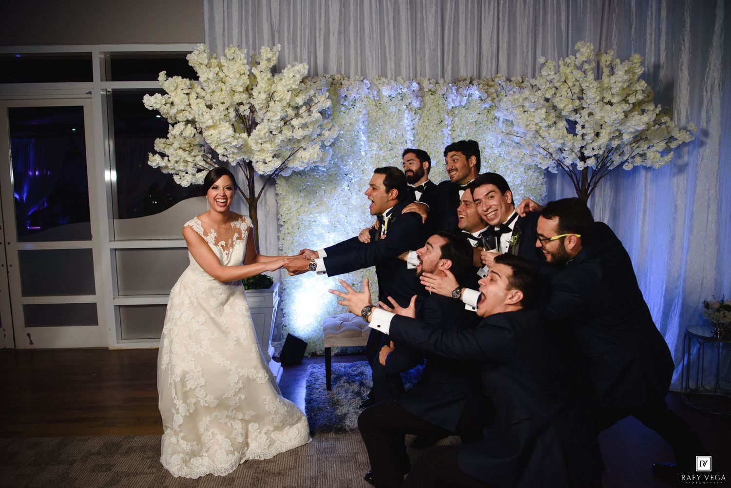Bride in white gown laughing with groomsmen in black suits near white floral trees at elegant wedding reception.
