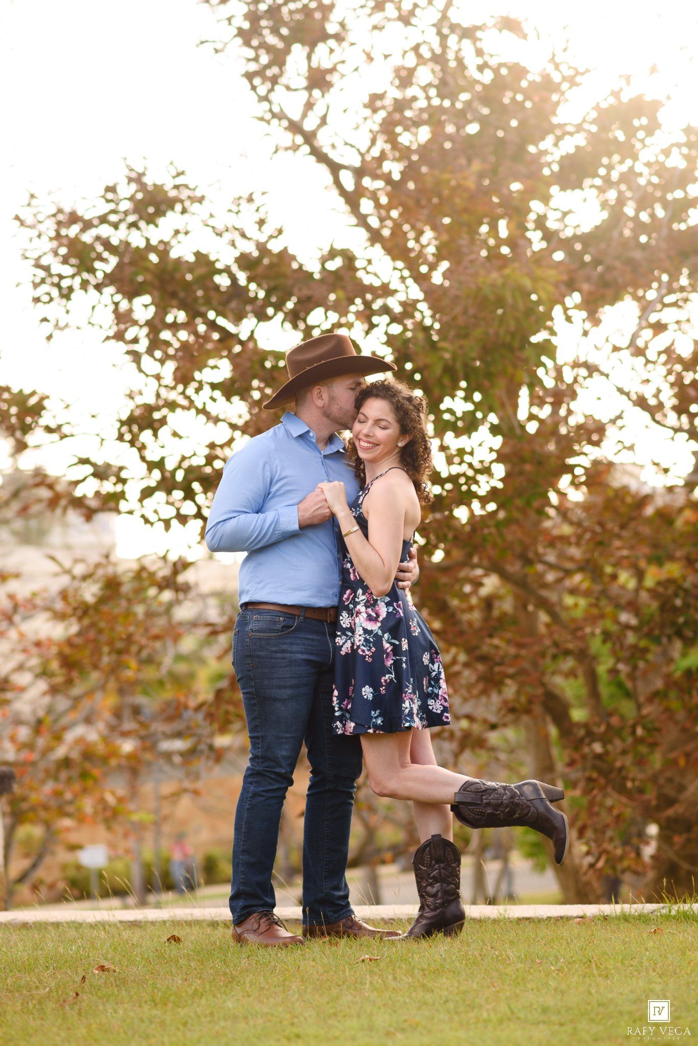 Couple embracing outdoors, man in cowboy hat and blue shirt kissing woman in floral dress and cowboy boots.