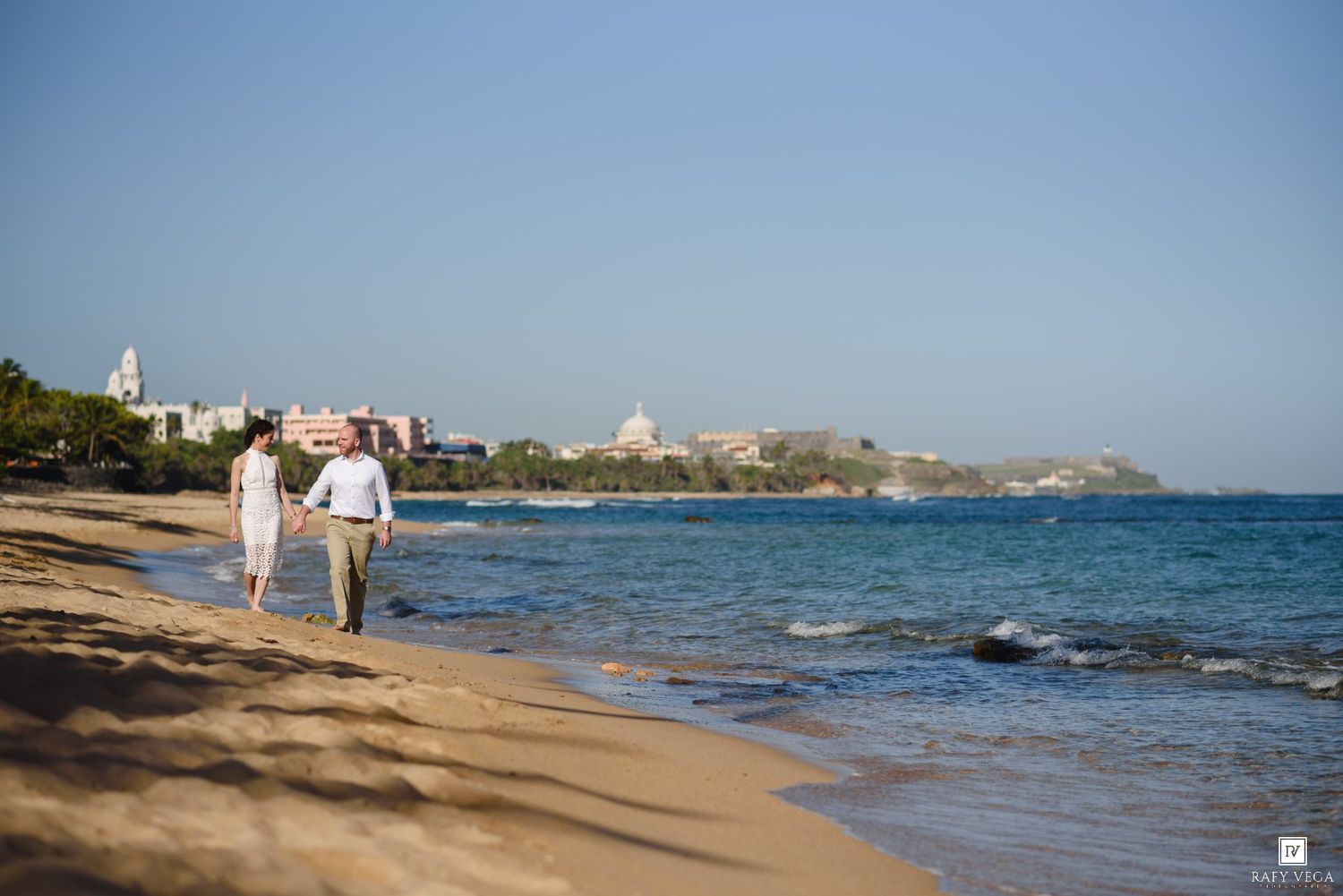 Couple walking hand in hand along a sandy beach with turquoise water and a historic cityscape in the background.